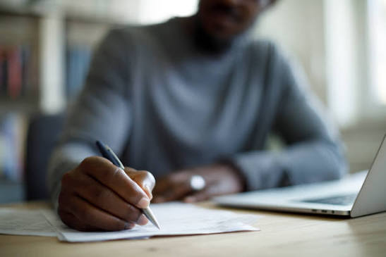 A focused photo of someone writing at a desk with a laptop or notebook